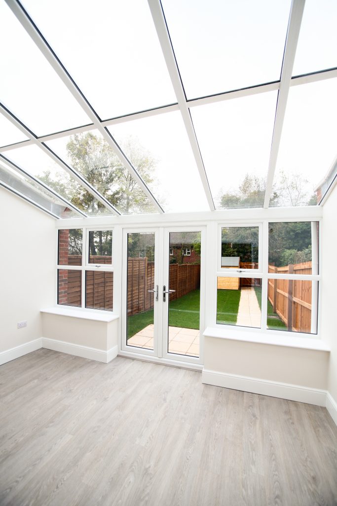 White interior of a lean-to conservatory with a glass roof and French doors leading to a garden.