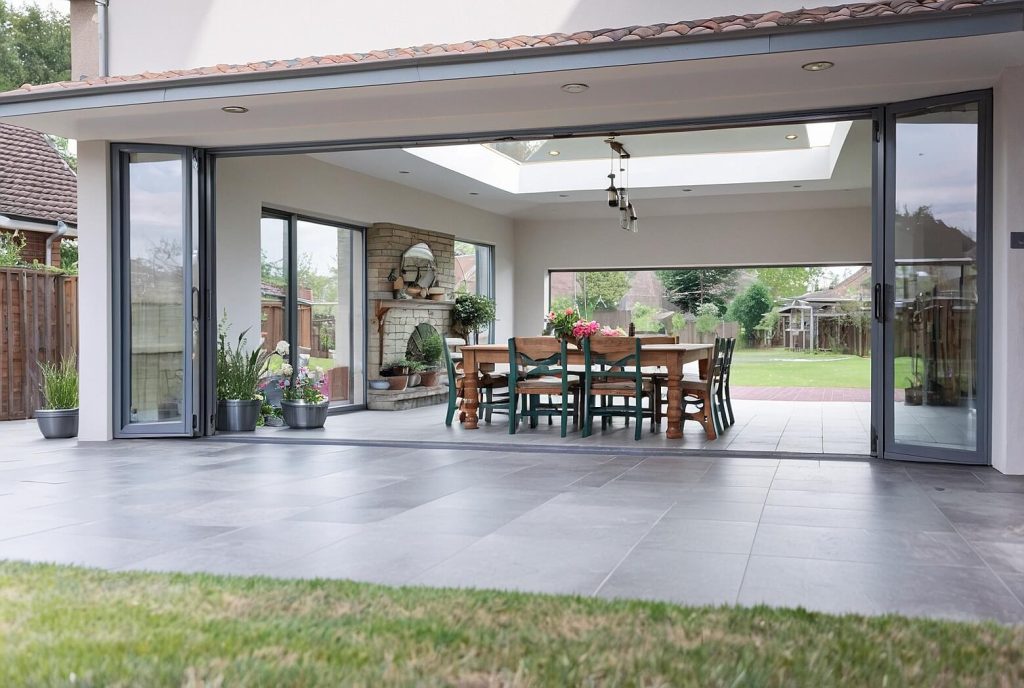 A contemporary home showcasing bifold doors that open fully to connect an indoor dining area with an outdoor patio. The dining space features a wooden table with chairs, plants, and a decorative fireplace, creating a seamless indoor-outdoor living experience.